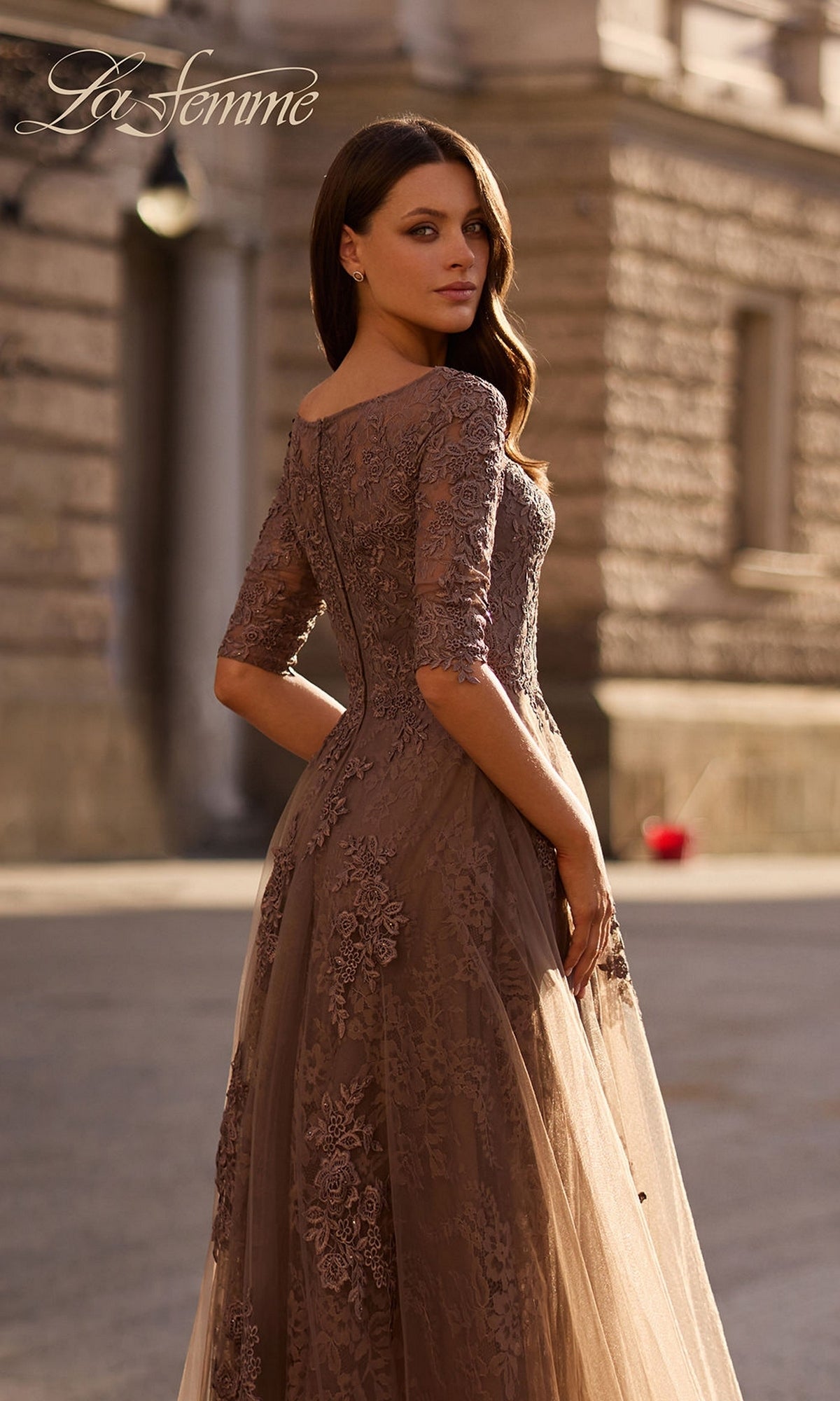 A woman in Long Formal Dress: La Femme 28036 stands outside, sunlight on her lace and tulle A-line skirt, with a blurred stone building behind. “La Femme” logo at top left.